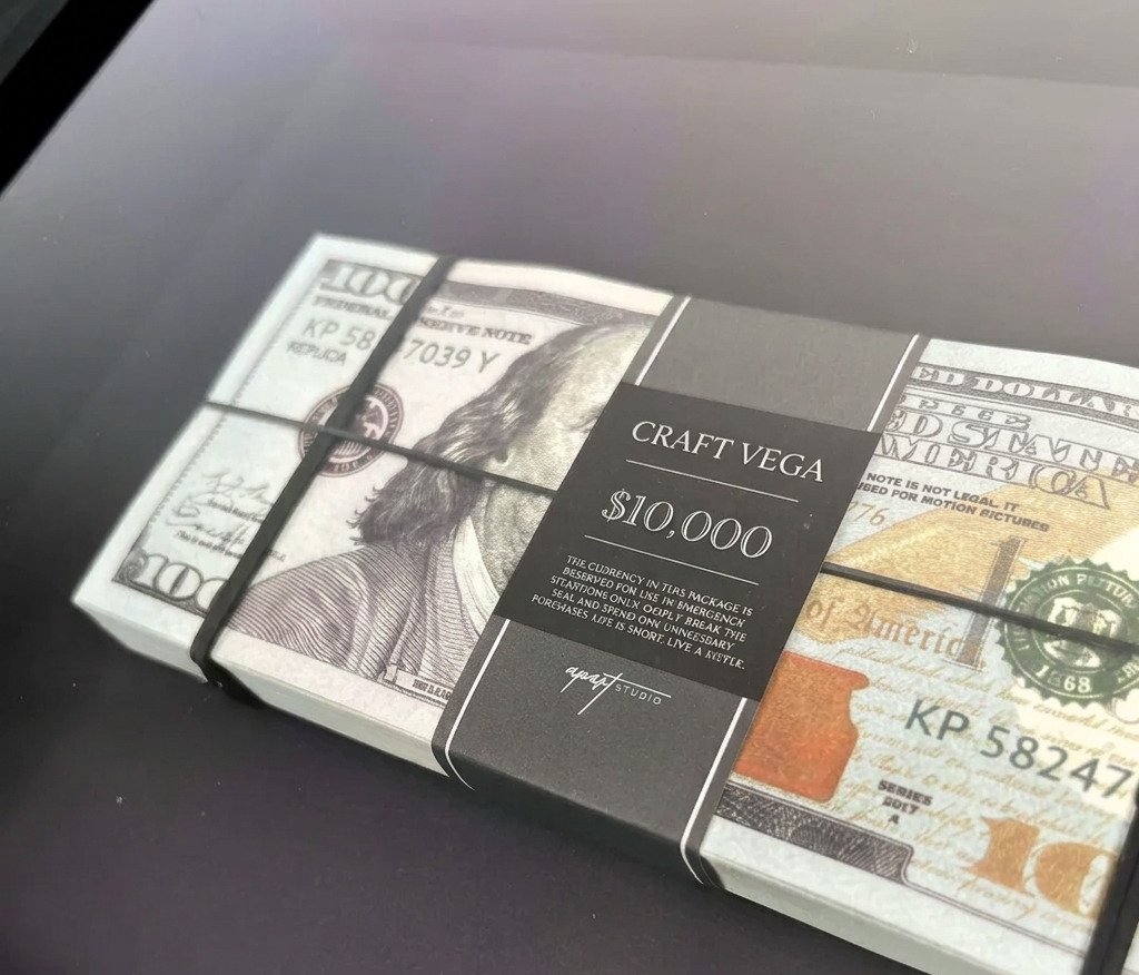 A stack of novelty prop money bundled with a black strap and label reading 'CRAFT VEGA $10,000' on a dark surface.